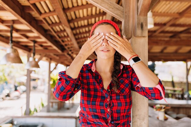 Bella donna caucasica con capelli scuri, pelle sana con trucco nudo pone alla macchina fotografica e viso chiuso con le mani in terrazza in legno