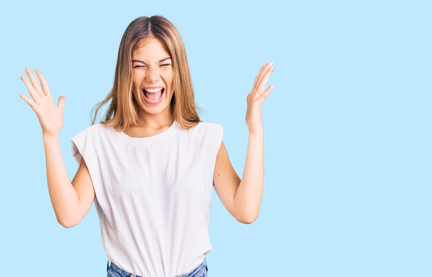 Bella donna caucasica con i capelli biondi che indossa una maglietta bianca casual che celebra la pazza e la pazza per il successo con le braccia alzate e gli occhi chiusi urlando il concetto di vincitore eccitato