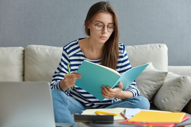 Bella donna caucasica in maglione a righe e occhiali, concentrata sui compiti, posa su un comodo divano in un appartamento moderno, utilizza il computer portatile per chattare online, posa a casa.