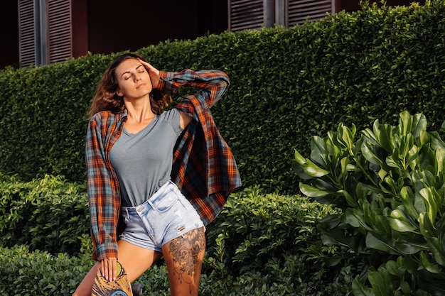 Bella donna caucasica tatuata in forma in pantaloncini di jeans, camicia a quadri con longboard