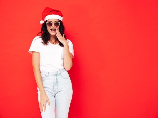 Bella donna che celebra il nuovo anno. Felice splendida femmina in abiti casual divertendosi alla festa di Capodanno. Celebrazione delle vacanze. Modello affascinante in Babbo Natale vicino al muro rosso. Scioccato e sorpreso