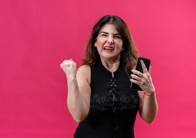 Bella donna che indossa camicetta nera guardando sorridente felicemente per la vittoria che tiene il telefono