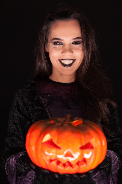 Bella donna che indossa un vestito da strega per halloween che tiene una zucca spettrale su sfondo nero.