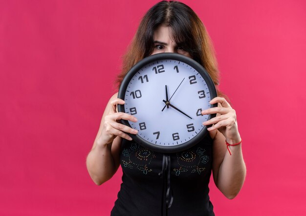 Bella donna che indossa una camicia nera che si nasconde dietro l'orologio su sfondo rosa