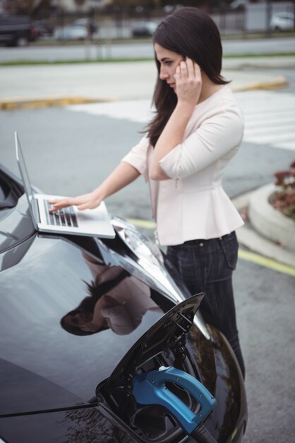 Bella donna che parla sul telefono cellulare durante la ricarica dell'auto elettrica