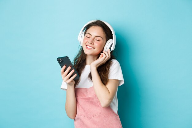 Bella donna che si gode la canzone in cuffia, chiude gli occhi e sorride mentre ascolta musica in cuffia, tenendo lo smartphone in mano, in piedi su sfondo blu.