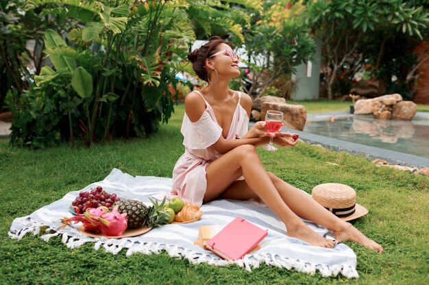 Bella donna che si siede sulla coperta, bevendo vino e godendo picnic estivo in giardino tropicale.