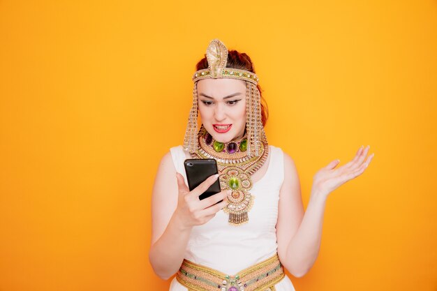 Bella donna come cleopatra in antico costume egiziano che tiene il braccio alzante dello smartphone con espressione delusa arrabbiata e frustrata sull'arancia