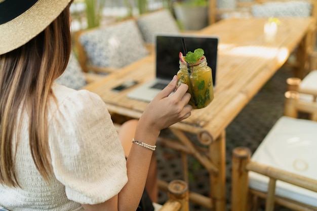 Bella donna con cappello di paglia che usa il lap top mentre viaggia in asia Lavora a distanza in un elegante caffè tropicale Tenendo bevande esotiche