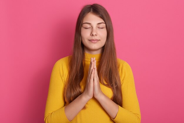 Bella donna con i capelli lunghi che prega con gli occhi chiusi, tenendo insieme il palmo, in posa isolato su uno spazio rosa, indossa un maglione giallo