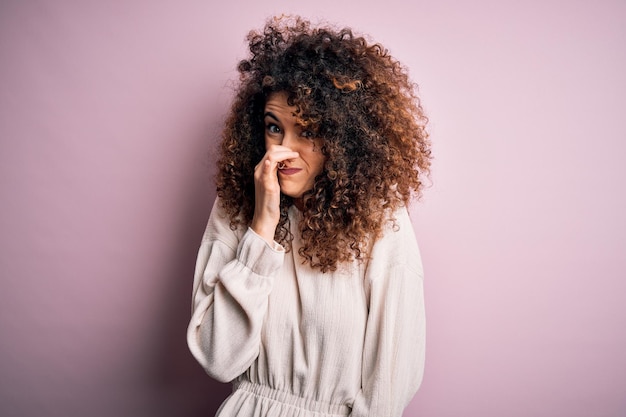 Bella donna con i capelli ricci e piercing che indossa un maglione casuale su uno sfondo rosa che puzza qualcosa di puzzolente e disgustoso puzzo insopportabile che tiene il respiro con le dita sul naso puzzo cattivo