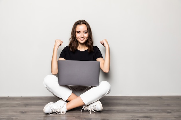 Bella donna con laptop moderno seduto sul pavimento con il gesto di vittoria sul muro grigio