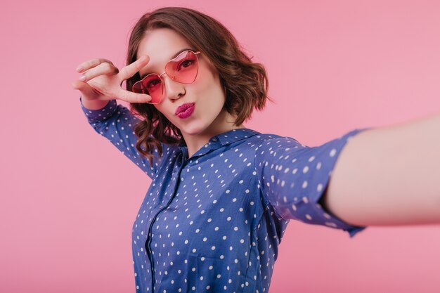 Bella donna con trucco luminoso che fa selfie con l'espressione del viso baciante sulla parete pastello. Foto di blithesome ragazza caucasica in occhiali da sole rosa in posa con il segno di pace.