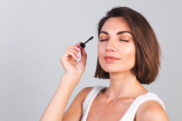 Bella donna con trucco tenere pennello mascara nero sul muro grigio, concetto di bellezza,