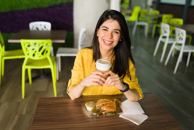 Bella donna con un sorriso raggiante che tiene un caffè e sta per iniziare a mangiare un gustoso panino sano presso la food court