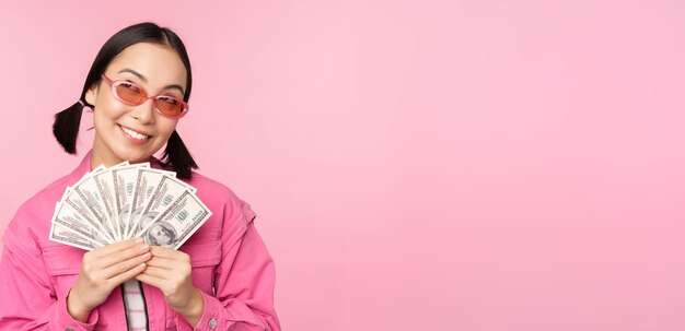 Bella donna coreana in occhiali da sole che mostra dollari denaro contante sorridente concetto soddisfatto di microcredito prestiti veloci e pagamento in piedi su sfondo rosa