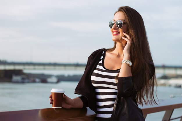 Bella donna d&#39;affari casuale sul suo telefono, ragazza con caffè al tramonto del fiume