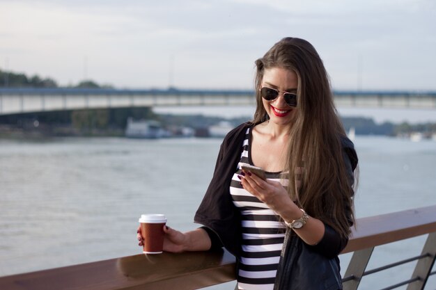 Bella donna d&#39;affari casuale sul suo telefono, ragazza con caffè al tramonto del fiume