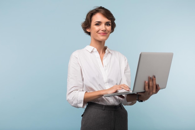 Bella donna d'affari sorridente con i capelli corti scuri in camicia bianca che tiene il laptop in mano mentre guarda felicemente nella fotocamera su sfondo blu