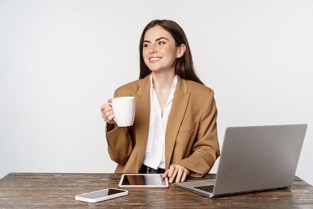 Bella donna d'ufficio felice, donna d'affari che beve caffè al lavoro, seduta al tavolo e sorride soddisfatta, con in mano una tazza bianca, sfondo dello studio.