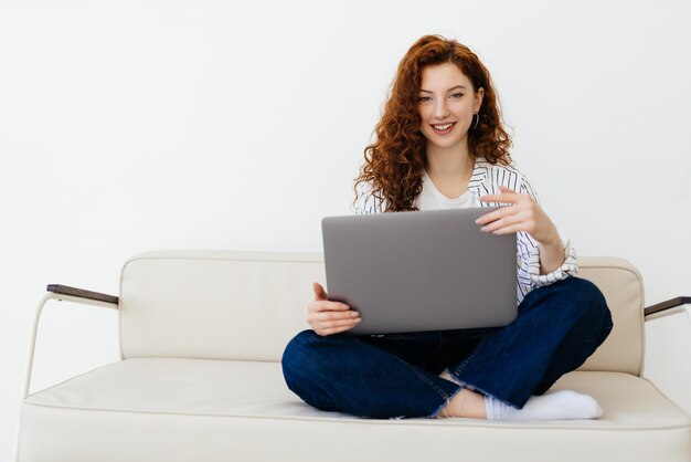 Bella donna dai capelli rossi che lavora online utilizzando il laptop seduto su un comodo divano grigio