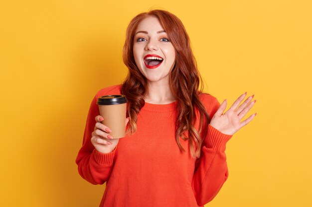 Bella donna dai capelli rossi preferisce il caffè da asporto, tiene la tazza usa e getta con una bevanda calda aromatica, guardando la telecamera con espressione felice e sorriso a trentadue denti