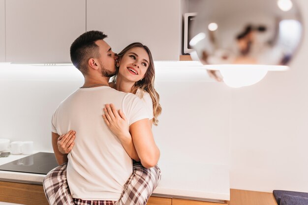 Bella donna dai capelli scuri divertendosi con il marito in cucina. Modello maschio brunetta in maglietta bianca in posa con la ragazza prima di colazione.