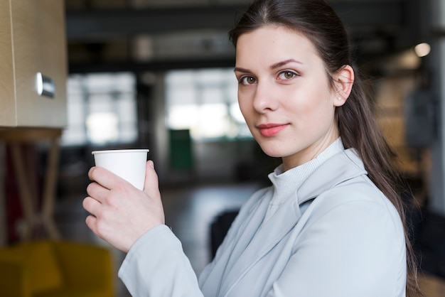 Bella donna di affari che tiene la tazza di caffè eliminabile che esamina macchina fotografica