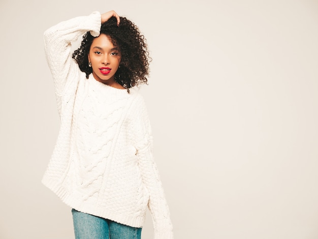 Bella donna di colore con l'acconciatura di riccioli afro e labbra rosse. Modello sorridente in vestiti di jeans alla moda e maglione invernale.