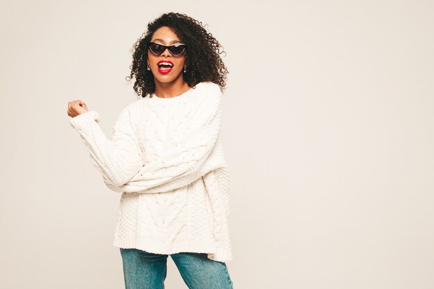 Bella donna di colore con l'acconciatura di riccioli afro e labbra rosse. Modello sorridente in vestiti di jeans alla moda e maglione invernale.