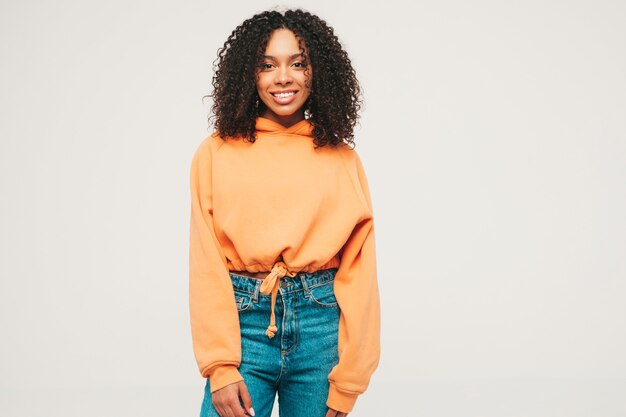 Bella donna di colore con l'acconciatura di riccioli afro. Modello sorridente in felpa con cappuccio arancione e vestiti di jeans alla moda