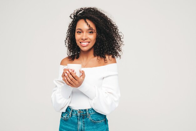 Bella donna di colore con l'acconciatura di riccioli afro. Modello sorridente in maglione e vestiti di jeans alla moda