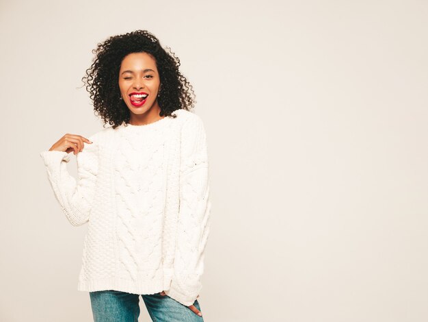 Bella donna di colore con l'acconciatura di riccioli afro. Modello sorridente in vestiti di jeans e maglione invernale bianco.