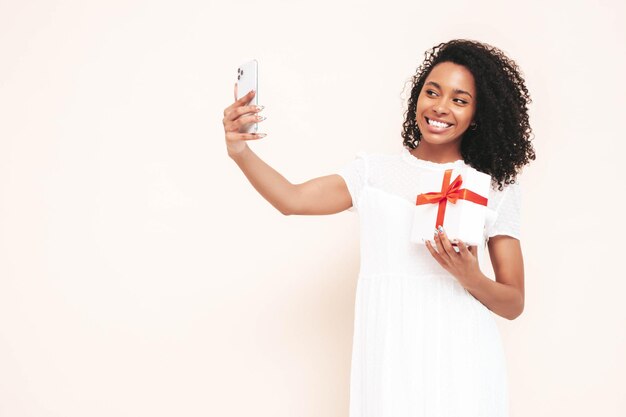 Bella donna di colore con l'acconciatura di riccioli afro Modello sorridente vestito in abito estivo bianco Femmina spensierata sexy in posa vicino al muro bianco in studio Scatola regalo abbronzata e allegra Holding Selfie