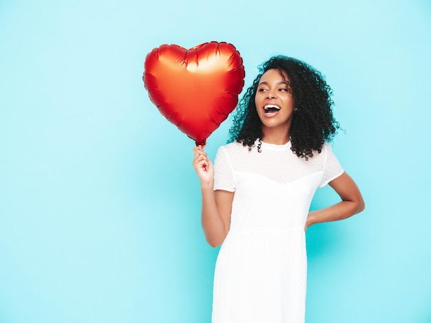 Bella donna di colore con l'acconciatura di riccioli afro Modello sorridente vestito in abito estivo bianco Femmina spensierata sexy in posa vicino al muro blu in studio Abbronzato e allegro Holding cuore mongolfiera