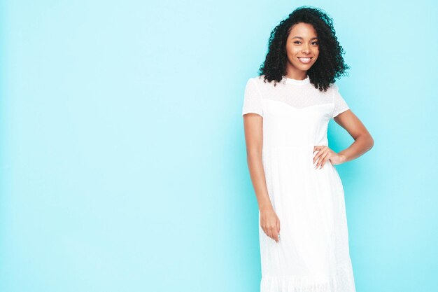 Bella donna di colore con l'acconciatura di riccioli afro Modello sorridente vestito in abito estivo bianco Femmina spensierata sexy in posa vicino al muro blu in studio Abbronzato e allegro Isolato