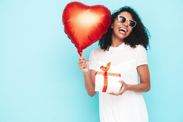 Bella donna di colore con l'acconciatura di riccioli afro Modello sorridente vestito in abito estivo bianco Femmina spensierata sexy in posa vicino al muro blu in studio Holding cuore mongolfiera e confezione regalo Isolato