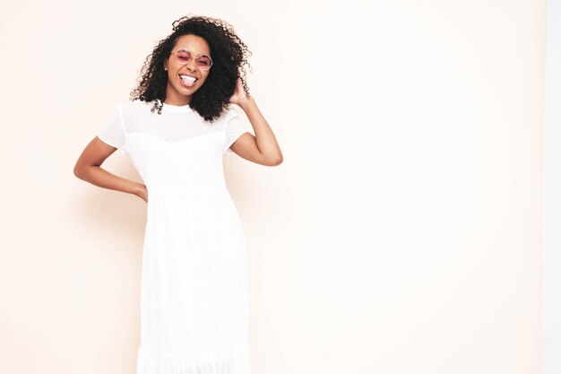 Bella donna di colore con l'acconciatura di riccioli afro Modello sorridente vestito in abito estivo bianco Femmina spensierata sexy in posa vicino al muro in studio Abbronzato e allegro Isolato Mostra la lingua