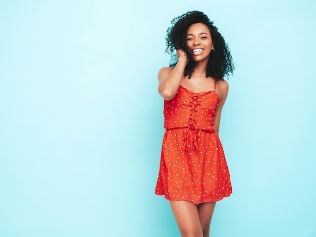 Bella donna di colore con l'acconciatura di riccioli afro Modello sorridente vestito in abito estivo rosso Femmina spensierata sexy in posa vicino al muro blu in studio Abbronzato e allegro Al giorno di sole isolato