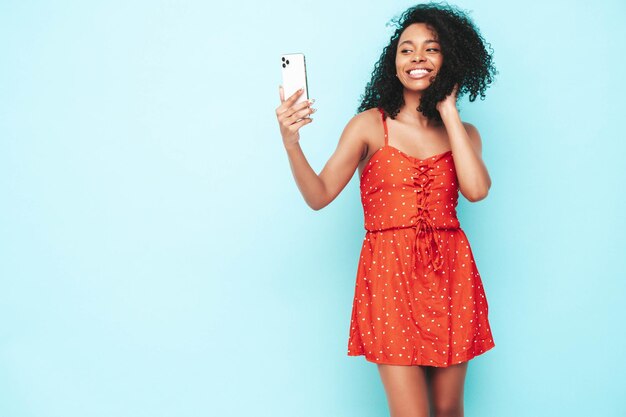 Bella donna di colore con l'acconciatura di riccioli afro Modello sorridente vestito in abito estivo rosso Femmina spensierata sexy in posa vicino al muro blu in studio Selfie abbronzato e allegro che prende