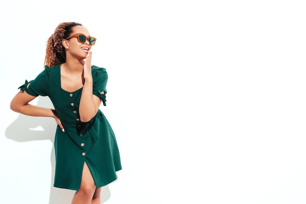 Bella donna di colore con l'acconciatura di riccioli afro Modello sorridente vestito in abito verde estivo bianco Femmina spensierata sexy in posa vicino al muro in studio Abbronzato e allegro isolato in occhiali da sole