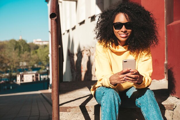 Bella donna di colore con l'acconciatura di riccioli afroModello hipster sorridente in felpa con cappuccio gialla Femmina spensierata sexy in posa sullo sfondo della strada in occhiali da sole Allegra e felice