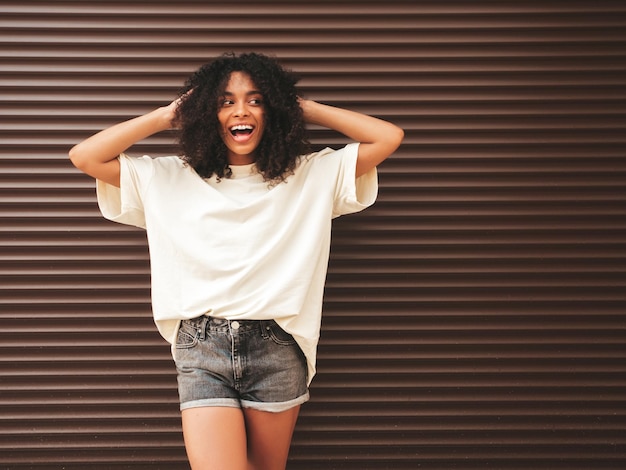 Bella donna di colore con l'acconciatura di riccioli afroModello hipster sorridente in maglietta bianca Femmina spensierata sexy in posa in strada vicino al muro marrone Allegro e felice in occhiali da sole