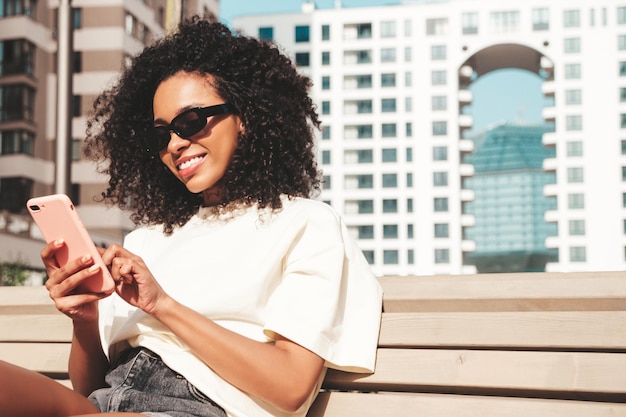 Bella donna di colore con l'acconciatura di riccioli afroModello sorridente in felpa con cappuccio bianca Donna sexy spensierata in posa sullo sfondo della strada in occhiali da sole Guardando lo schermo dello smartphone utilizzando le app