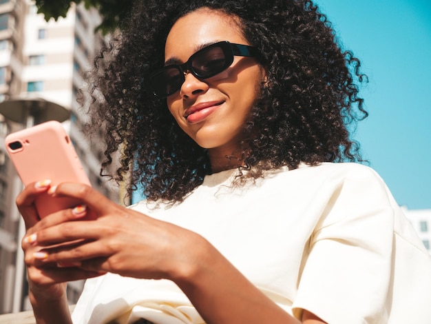 Bella donna di colore con l'acconciatura di riccioli afroModello sorridente in felpa con cappuccio bianca Donna sexy spensierata in posa sullo sfondo della strada in occhiali da sole Guardando lo schermo dello smartphone utilizzando le app