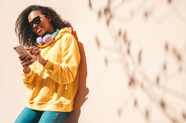 Bella donna di colore con l'acconciatura di riccioli afroModello sorridente in felpa con cappuccio gialla Donna sexy spensierata in posa sullo sfondo della strada in occhiali da sole Guardando lo schermo dello smartphone utilizzando le app