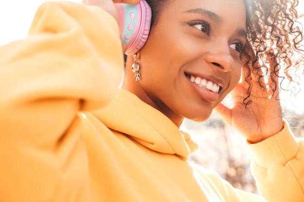 Bella donna di colore con l'acconciatura di riccioli afroModello sorridente in felpa con cappuccio giallaFemmina sexy spensierata che si gode l'ascolto di musica in cuffie wireless Posa su sfondo di strada al tramonto