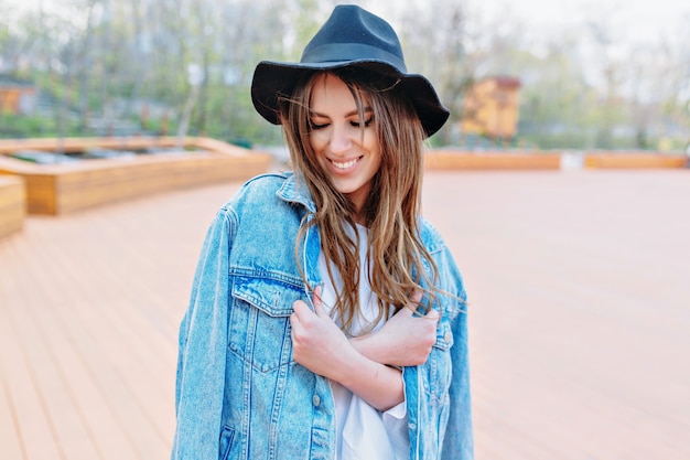 Bella donna di stile con lunghi capelli scuri che indossa un cappello nero e giacca di jeans chiude gli occhi con un sorriso splendido. Atmosfera estiva. Ritratto all'aperto nel parco