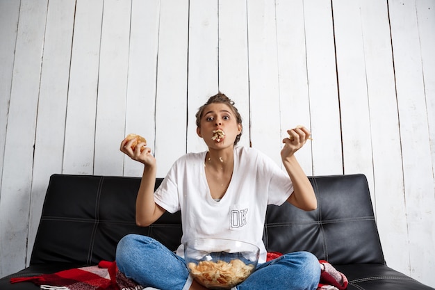 Bella donna domestica mangiare patatine, guardare la tv, seduto al divano.