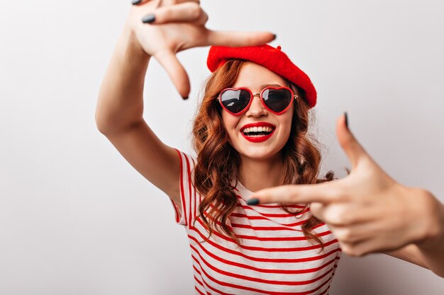 Bella donna europea in berretto rosso che esprime felicità. Ragazza riccia emozionante in occhiali da sole che ride.